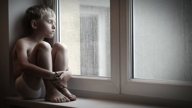 Moody little boy sitting on the windowsill watching raindrops go down the window