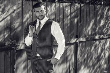 Black-white portrait of young beautiful fashionable man against wooden fence In classic suit.

