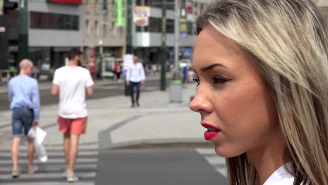 young attractive blonde woman looks around - urban street in the city with cars and people in the background - pedestrian crossing - closeup face