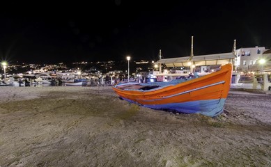 Fototapeta premium Night view of Chora port in Mykonos, Greece. Hora town cityscape lights reflected on the sea, a typical orange, blue greek island fishing boat in the harbour sandy shore and whitewashed buildings.