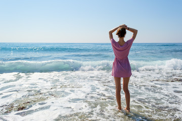 Young woman in summer dress standing by blue sea and looking to horizon