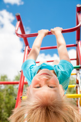 Obraz premium Low angle view of cute blond girl wearing blue teeshirt hanging from a monkey bars. Girl is hanging upside down looking at camera smiling