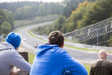A group of friends watching a car race on a legendary racing track in Germany. People are watching the race behind a metal fence.