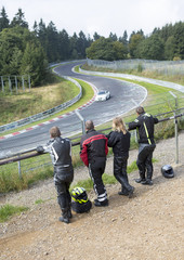 A group of friends watching a car race on a legendary racing track in Germany. People are watching the race behind a metal fence.