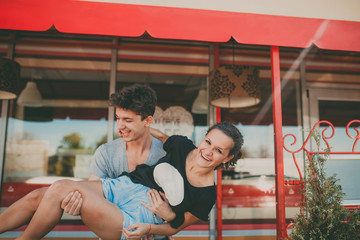 Couple in love having fun outside a cafe. Handsome young man holding his girlfriend on hands. Young teenage people laughing