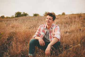 Handsome teenage boy wearing casual shirt sitting on grass on autumn day. Happy smiling young man...