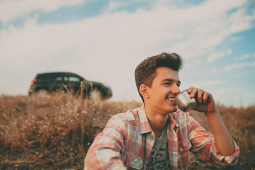 Confident smiling teenage boy sitting outdoors by his suv car on background and drinking tea