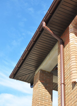 New Rain Gutter Drain Pipe Downspout Installation on the Unfinished House Facade Brick Wall Outdoor. Install Drainage System with Plastic Siding Soffits and Eaves Exterior.