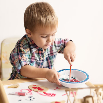 Adorable kid playing on his table with watercolors, painting a wooden parts of his airplane wooden toy. He is young designer with many ideas waiting to be found. Shallow depth of field.
