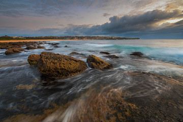 Fototapeta na wymiar Seascape sunrise long exposure with flowing water around large rocks and light pink and blue sky