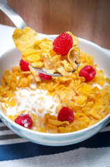 Bowl of corn flakes and raspberries on a table cloth in a rustic kitchen with a spoon scooping some cereal and a raspberry