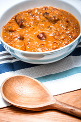 Bowl of delicious homemade beans and sausage in a rustic environment, on a wooden table and a table cloth with a wooden spoon in the frame.