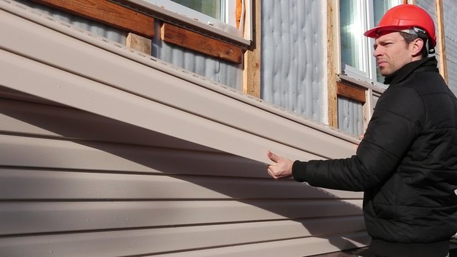 A worker installs panels beige siding on the facade of the house
