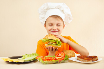 Little funny chef in chefs hat enjoys a cooking tasty hamburger