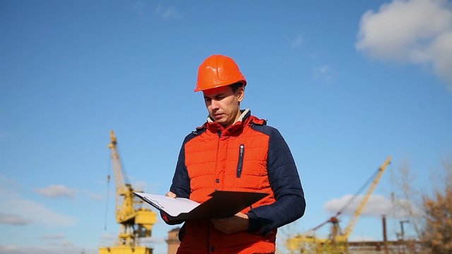 man construction worker in an orange helmet holding documents in the background blue sky and a crane
