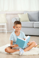Little boy with book sitting on carpet, on home interior background