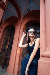 young beautiful brunette girl in a hat, sunglasses, t-shirt and jeans, with a film camera walks in the old city, holding hand hat for fear of losing it