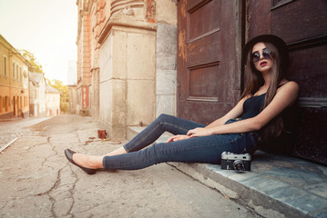 young beautiful brunette girl in a hat, sunglasses, t-shirt and jeans, with a film camera walks in the old city, holding hand hat for fear of losing it - obrazy, fototapety, plakaty