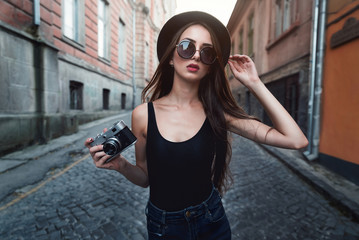 young beautiful brunette girl in a hat, sunglasses, t-shirt and jeans, with a film camera walks in the old city, holding hand hat for fear of losing it