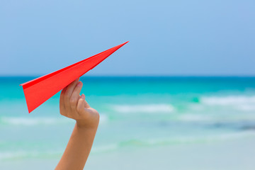 Female hand playing with red paper plane on the beach on blue sea background