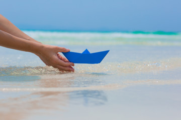 Female hands playing with blue paper boat in water on the white sand beach on blue sea background