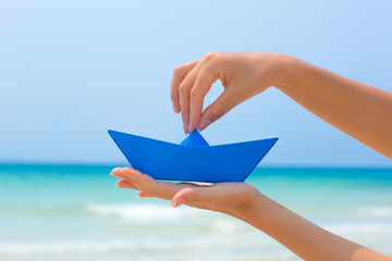 Female hands playing with blue paper boat in water on the white sand beach on blue sea background