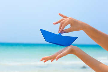 Female hands playing with blue paper boat in water on the white sand beach on blue sea background
