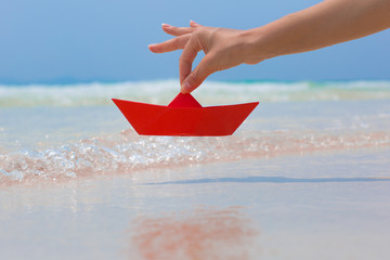 Female hand playing with red paper boat in water on the white sand beach on blue sea background