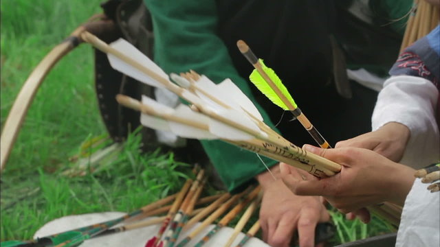 Medieval archers prepare arrows, sitting on the grass.