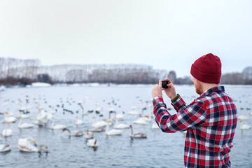 man in shirt and hat photographing swans winter