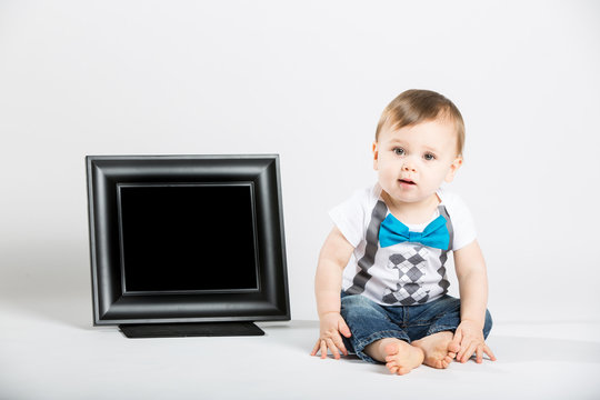 a cute 1 year old baby sits next to a blank black picture frame in a white studio setting. The boy is looking at the camera curiously. He is dressed in Tshirt, jeans, suspenders and blue bow tie