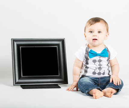 a cute 1 year old baby sits next to a blank black picture frame in a white studio setting. The boy is sitting in inticipation. He is dressed in Tshirt, jeans, suspenders and blue bow tie