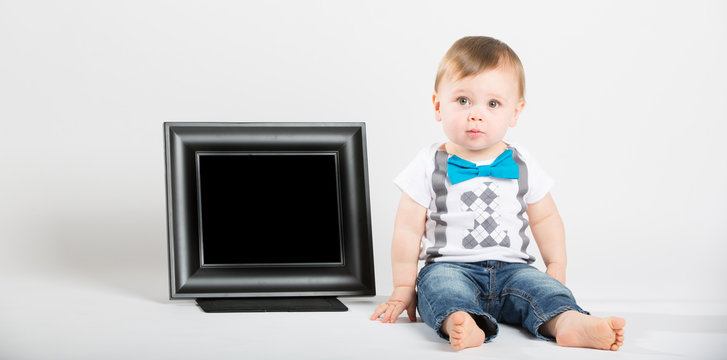 a cute 1 year old baby sits next to a blank black picture frame in a white studio setting. The boy has a confused expression. He is dressed in Tshirt, jeans, suspenders and blue bow tie