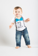 a cute 1 year old stands in a white studio setting. The boy has a happy expression. He is dressed in Tshirt, jeans, suspenders and blue bow tie
