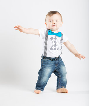 a cute 1 year old stands in a white studio setting. The boy looks as if he is surfing in the studio.. He is dressed in Tshirt, jeans, suspenders and blue bow tie