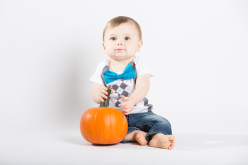Fototapeta premium a cute 1 year old sits in a white studio setting with a pumpkin. The boy looks up while holding the stem of the pumpkin. He is dressed in Tshirt, jeans, suspenders and blue bow tie