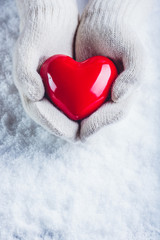 Fototapeta premium Female hands in white knitted mittens with a glossy red heart on a snow background. Love and St. Valentine concept.