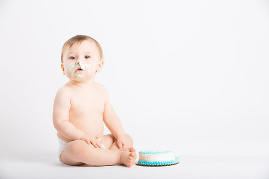 A Cute 1 Year Old Sits In A White Studio Setting. The Boy Looks Over With A Face Full Of Cake Icing For His Birthday. He Is Only Dressed In A White Diaper