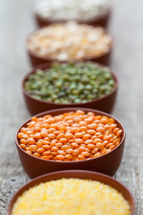 Bowls of cereal grains: red lentils, green mung, corn, beans and peas on wooden table. Selective focus on red lentils.