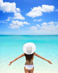 Fototapeta na wymiar Beach woman standing with arms outstretched against turquoise sea and blue sky. Rear view of female wearing white sunhat and bikini. Carefree tourist is enjoying vacation at beach.