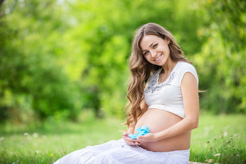 Naklejka na ściany i meble Beautiful pregnant woman holding baby booties and smiling outside in the park