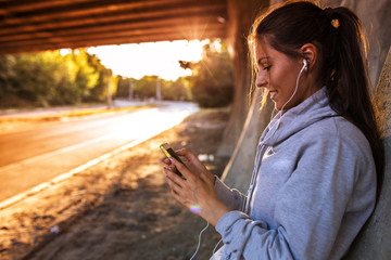 Young woman relaxing after jogging.She using her smart phone and listening to music.Typing message.leaning against wall. - obrazy, fototapety, plakaty