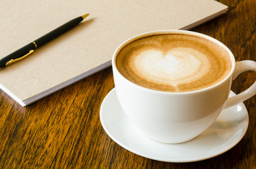 A cup of coffee with heart shape, pen and blank book on wood bac