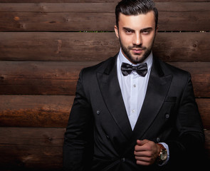 Portrait of young beautiful fashionable man against wooden wall In black suit & bow tie.
