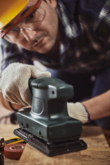 Close-up portrait of young handyman wearing protective helmet, glasses and gloves working with electric planer in workshop.  