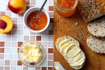 Tasty jam in the jar and bowl, ripe peaches, bread with butter and wooden tablet on mosaic background