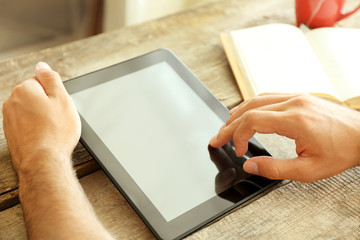 Tablet in man's hands, book and cup of tea on wooden table