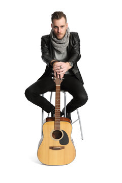 A man wearing a black leather jacket sitting down with an acoustic guitar. White background.