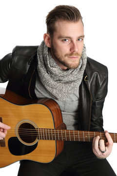 A man wearing a black leather jacket sitting down with an acoustic guitar. White background.