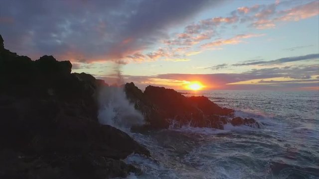 AERIAL: Waves splashing against rocky shore at golden sunset